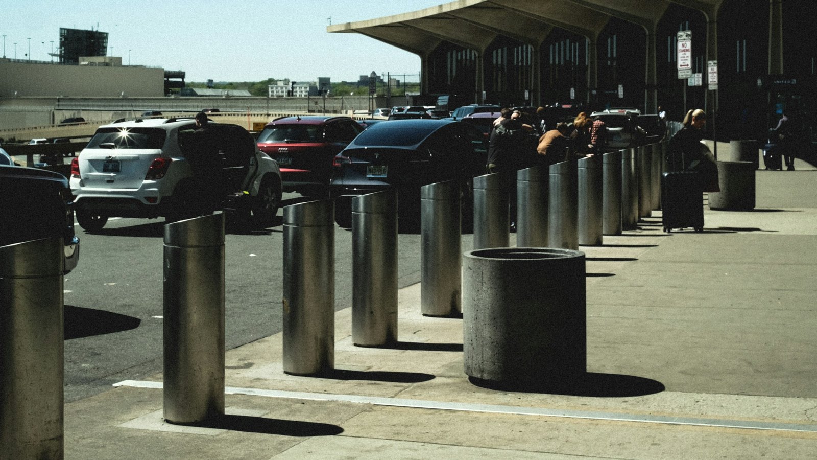 Kerbside pickup area under the Newark airport terminal canopy, with cars queued and travellers wheeling suitcases